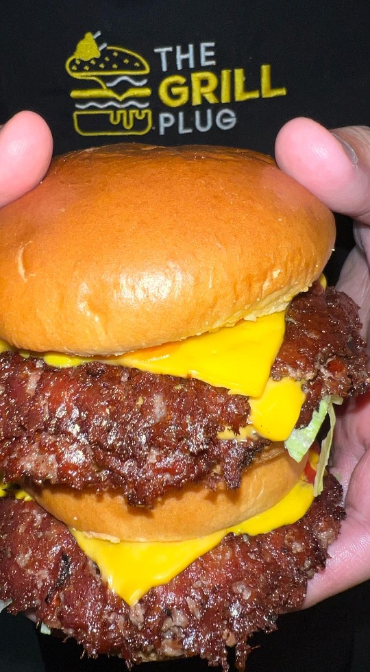 The Grill Plug, serving The Best Burgers! Delivery in Cambridge Close up of a smashburger being held by a person wearing a The Grill Plug t shirt